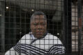 Erick Mauro Nguémah, derrière les grilles, à trois jours de la probable inauguration du monument qu'il a porté pendant trois ans. (Illustration générée par IA) © GabonReview